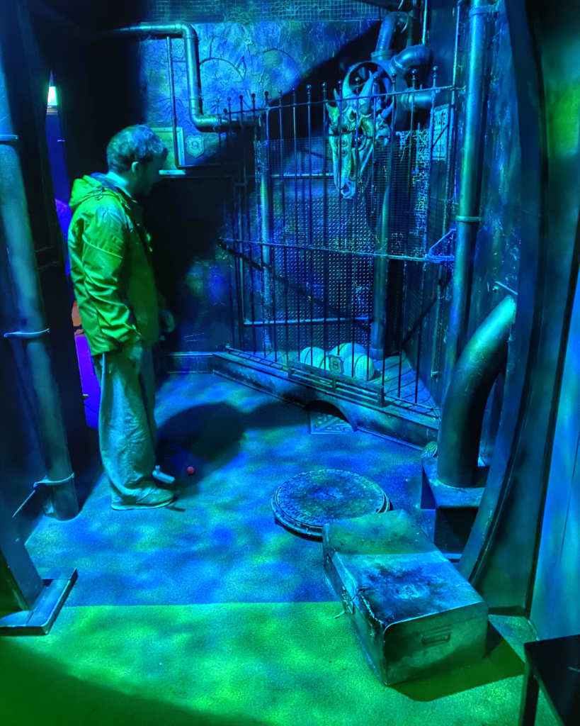 A man holding a golf club stans in front of a metal gate with a dragon's head behind it. The room is lit with dark green and blue light.