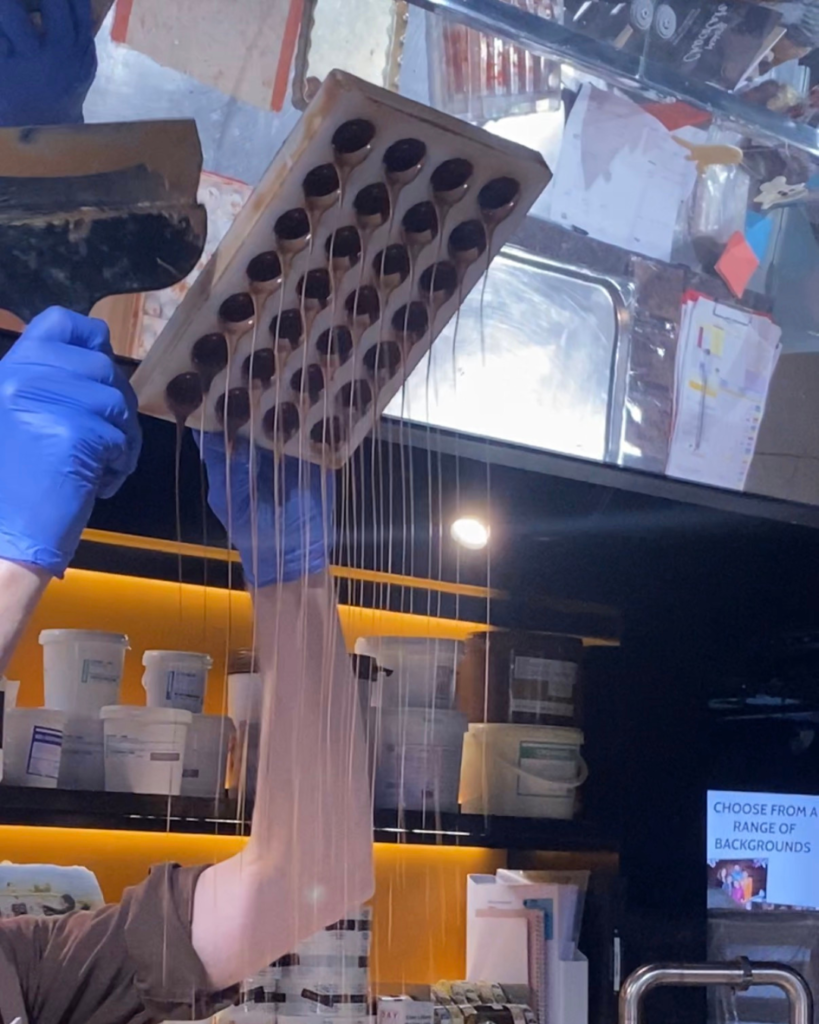 An arm holds a chocolate mold overhead, with strings of melted chocolate pouring downwards.