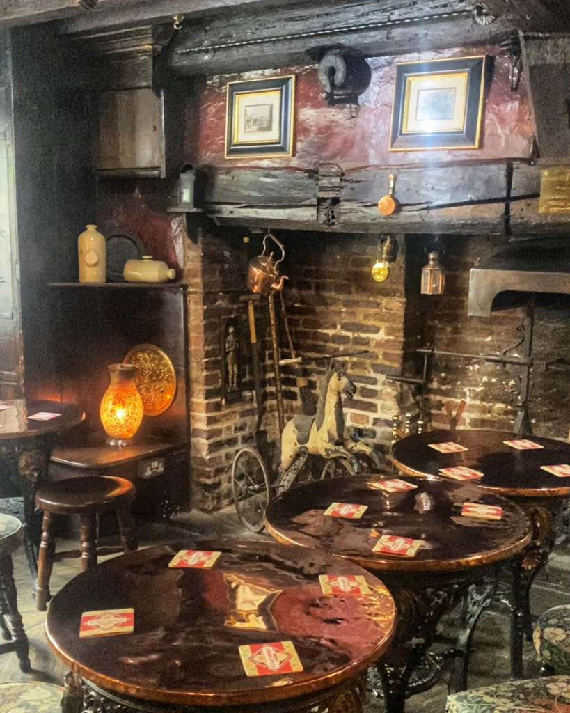 Pub tables with stools are in front of a brick fireplace with timber walls. An old rocking horse stands next to the fire.