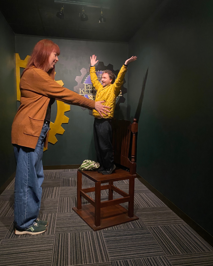 An optical illusion showing a tall woman holding a small man standing on a seat. 