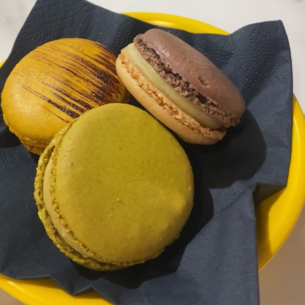 A yellow plate with three macarons: one a dark yellow, one green and one brown and cream.