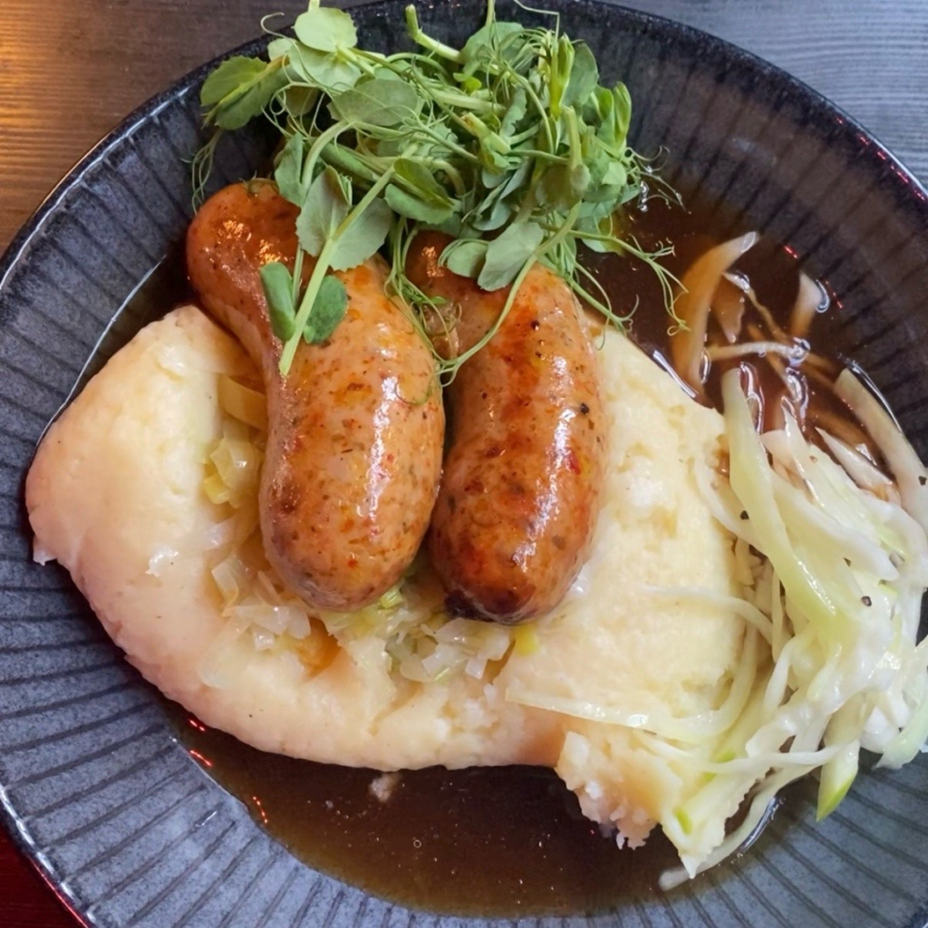 A close up of a plate showing two fat sausages on a bad of mash, with gravy and greens on the side. 