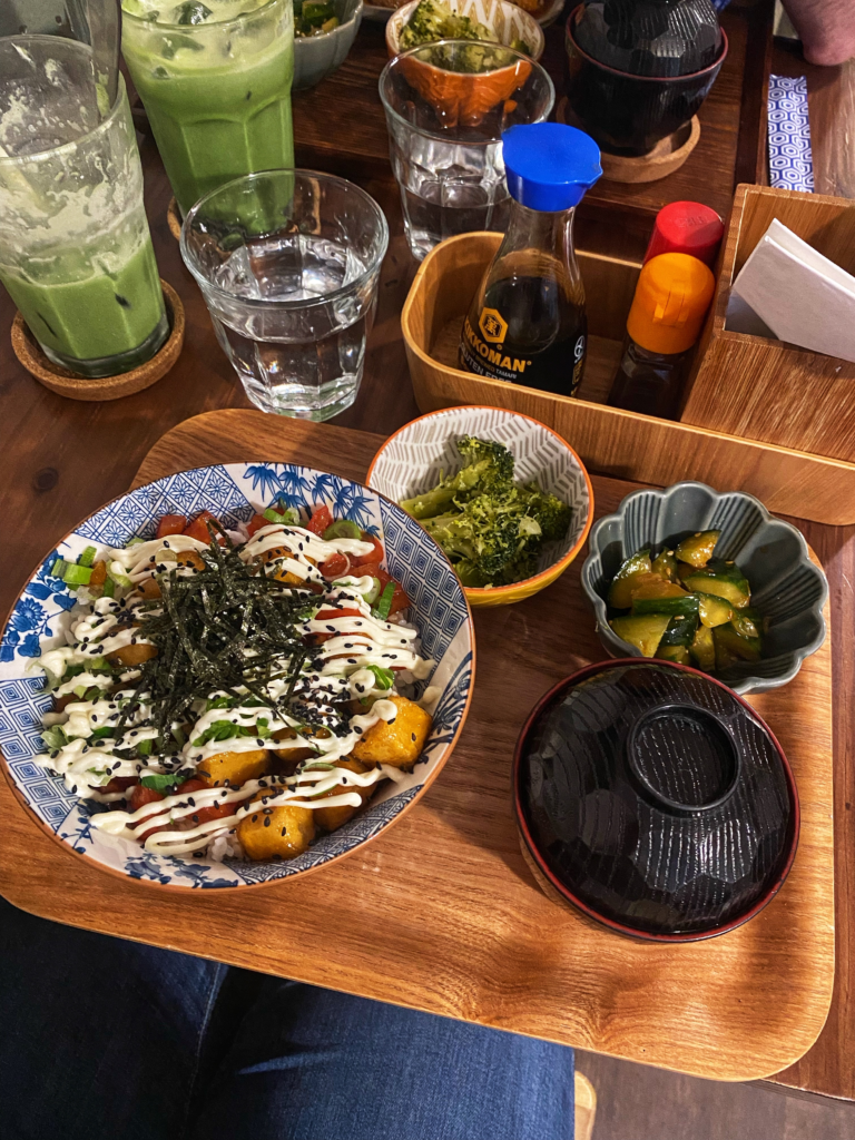 A table with a tray containing a bowl of tofu and vegetables, a smaller bowl of miso and two tiny bowls of vegetables. Two glasses of iced matcha are in the background.