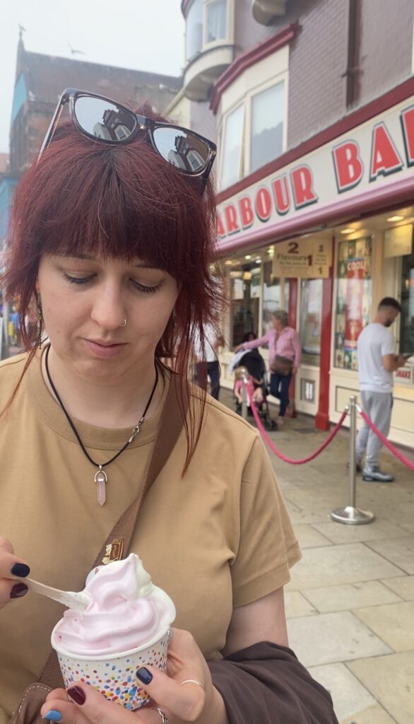 A woman looks down at a tub containing pink and white soft serve ice cream. A shop front with red letters can be seen behind.
