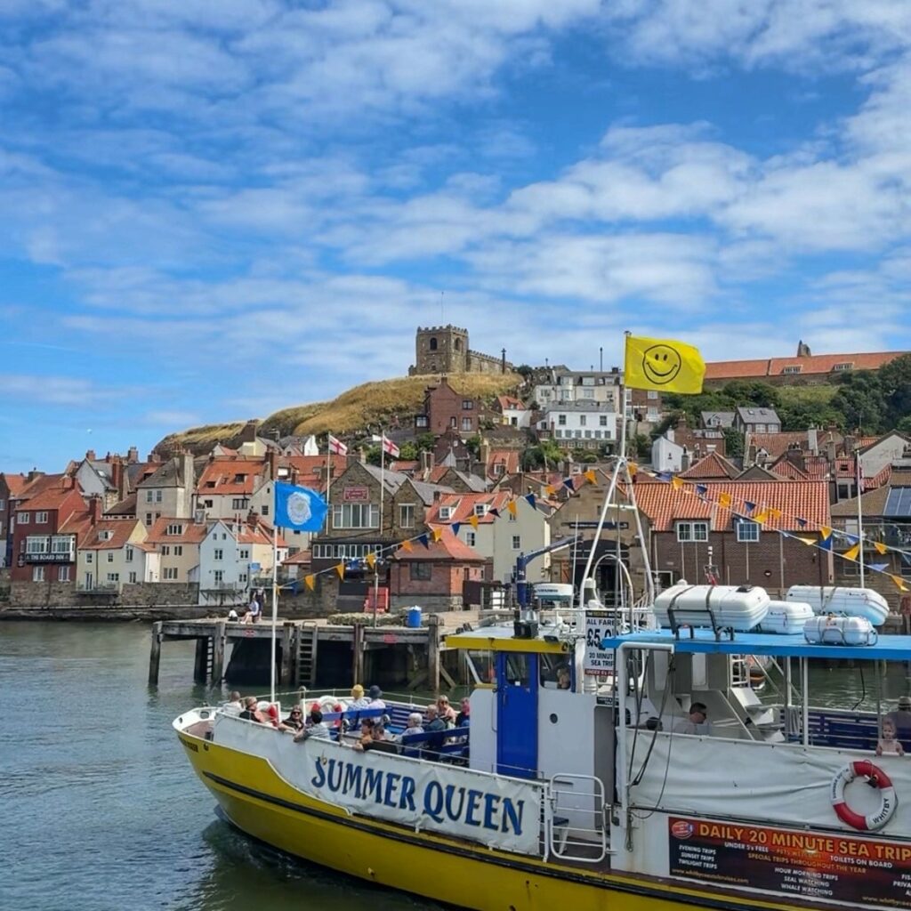 A white and yellow passenger boat sails past, with Whitby in the background and a church seen on the hill above