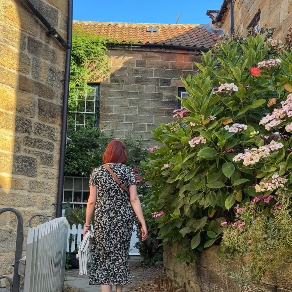 A woman in a black floral dress walks in a small passageways between stone cottages